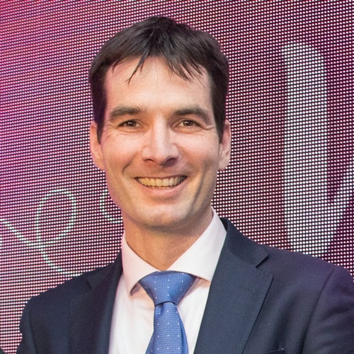 Portrait of a person in a suit and tie, smiling, against a purple background.