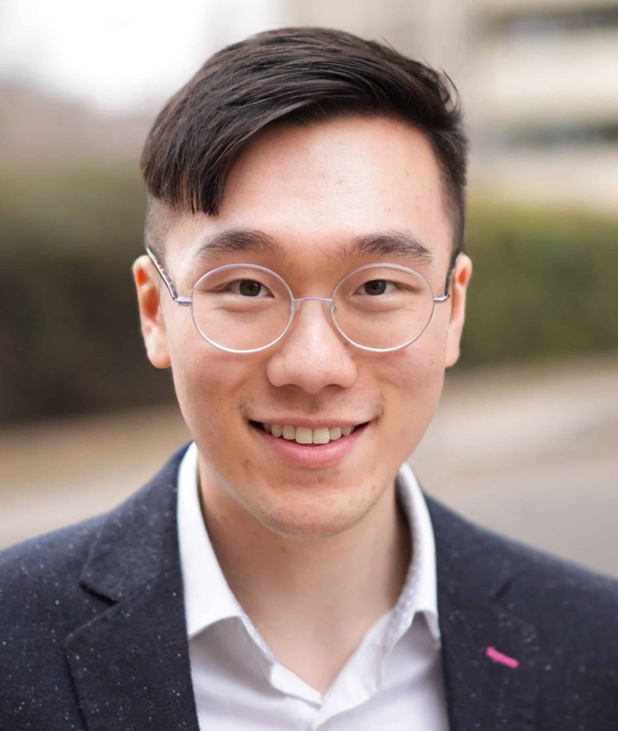 Head-and-shoulders portrait of a person wearing round glasses and a dark blazer over a light-colored shirt, standing outdoors with a softly blurred background. The person is positioned centrally in the frame and is facing the camera directly.