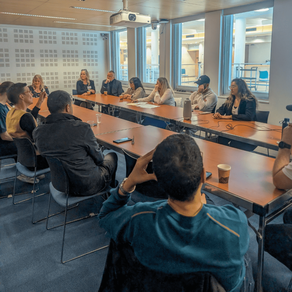 A wider group discussion is taking place around a large meeting table. A mix of participants, including men and women, are seated and engaged in a conversation. Natural light from tall windows brightens the room.