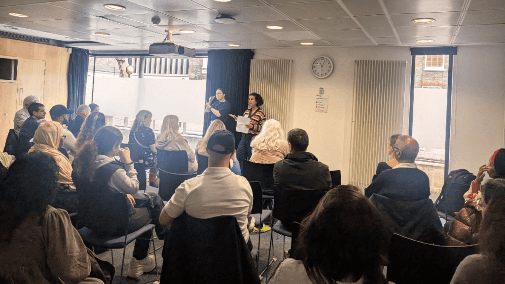 A woman presents to a seated audience in a meeting room, with a sign language interpreter standing beside her. Attendees are listening attentively. The event is taking place during the day with natural light coming through the windows.