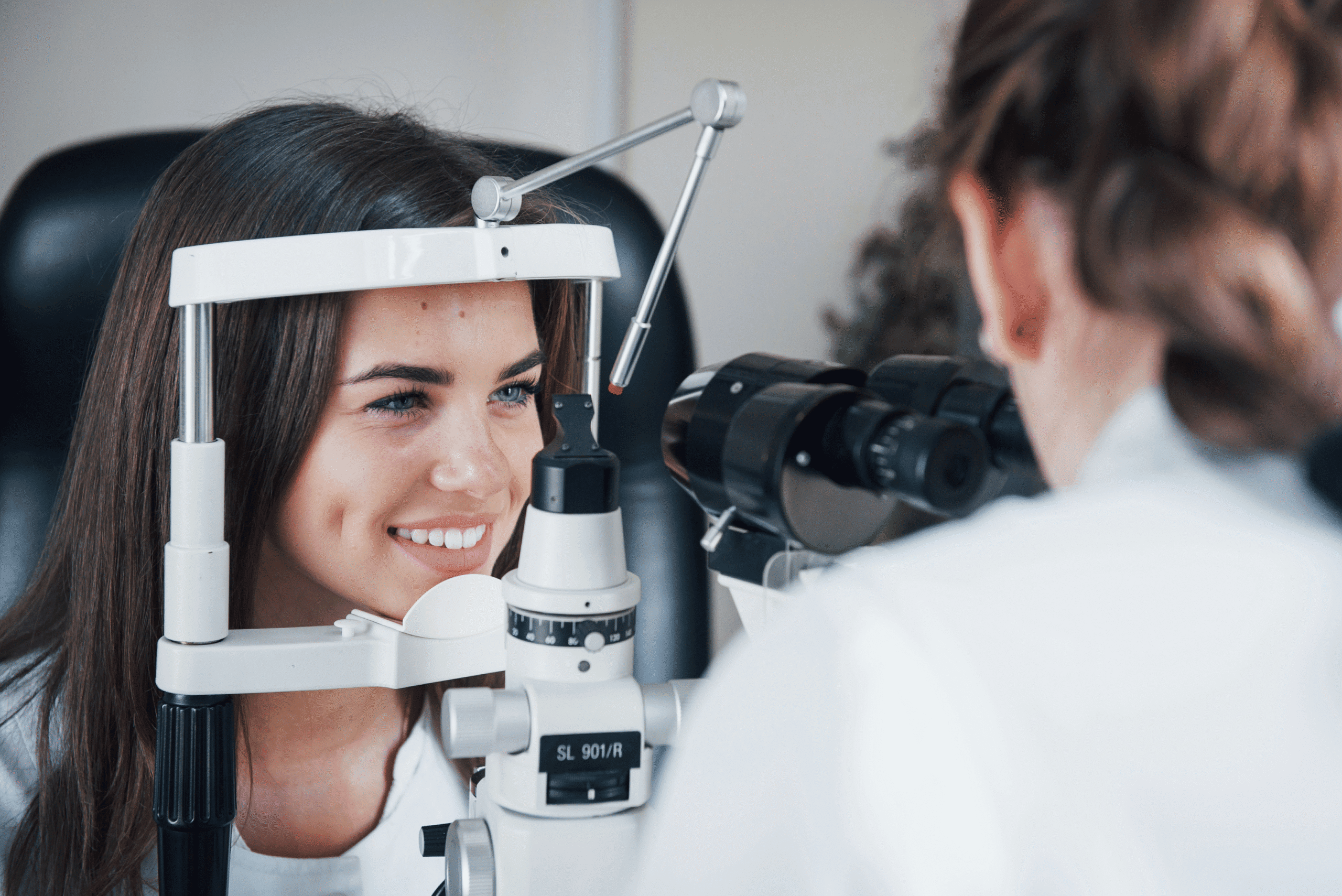 Image shows a young woman is undergoing an eye examination by an optometrist using a slit lamp. The woman is smiling and resting her chin on the device’s support as the optometrist looks through the instrument to inspect her eyes. The setting appears to be a modern optometry clinic.