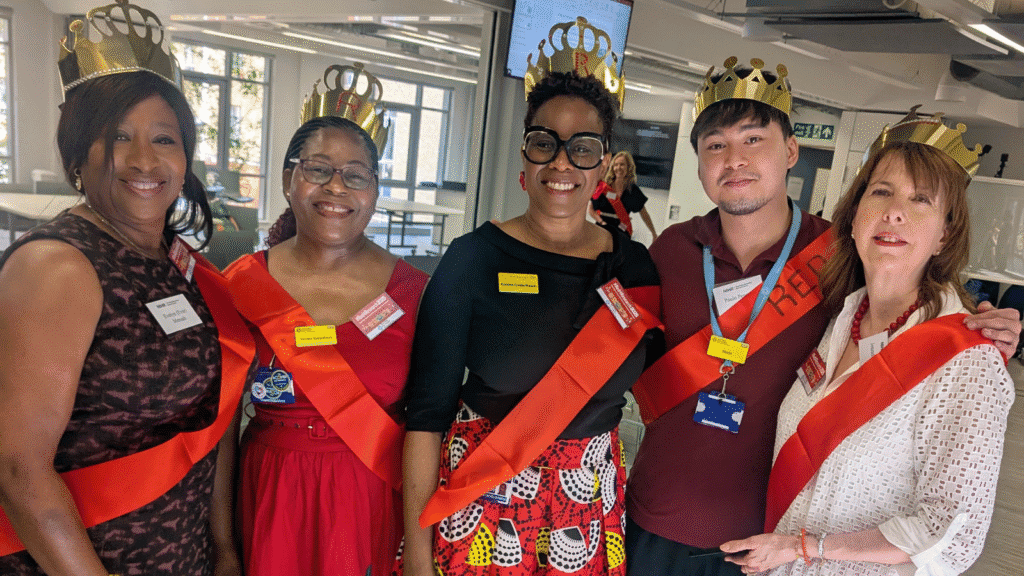 A group of five people standing together indoors, each wearing a gold crown and a red sash. They appear to be in a celebratory or formal event setting, with name tags visible on their clothing. The background shows a modern room with large windows and some furniture.