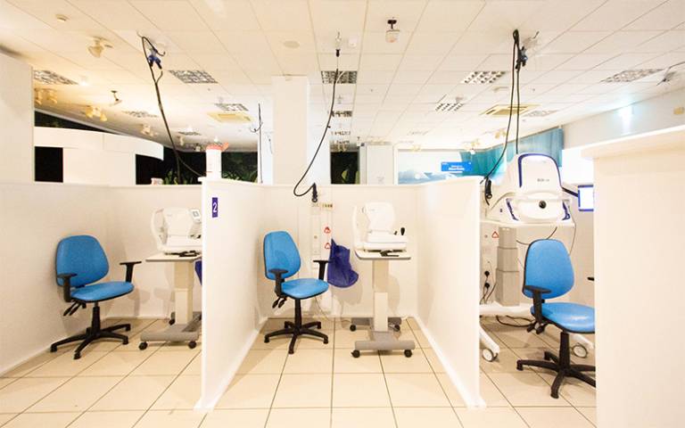 A clean, well-lit room with three cubicles, each containing an eye examination station. Each station includes a blue chair and white ophthalmic equipment. The cubicles are separated by white partitions, and cables hang from the ceiling connecting to the equipment. The room has tiled flooring and multiple ceiling light fixtures