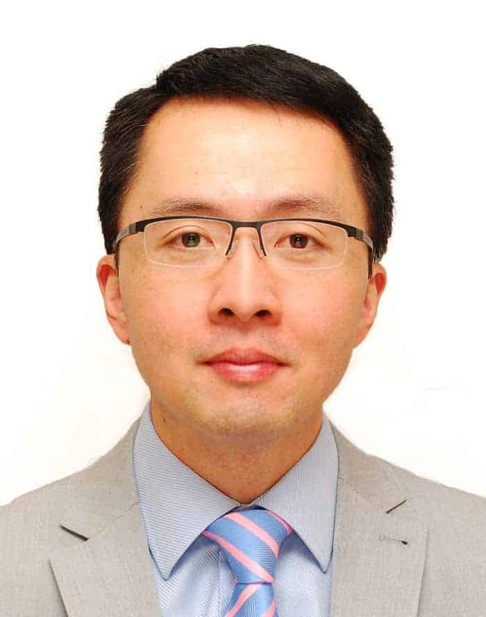 individual in grey suit and tie with black glasses looks straight into the camera