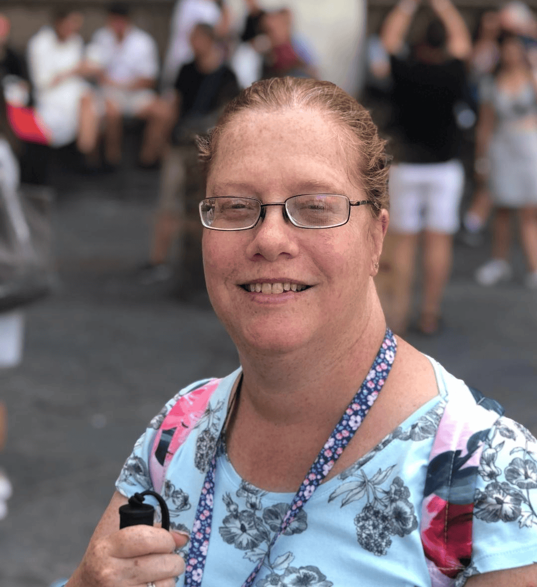 A person wearing a light blue floral-patterned top and a lanyard is standing outdoors in a busy public area. The individual is holding a black object in one hand. In the background, several people are visible, some sitting and others standing, with a mix of casual clothing and blurred surroundings suggesting a social or tourist setting.