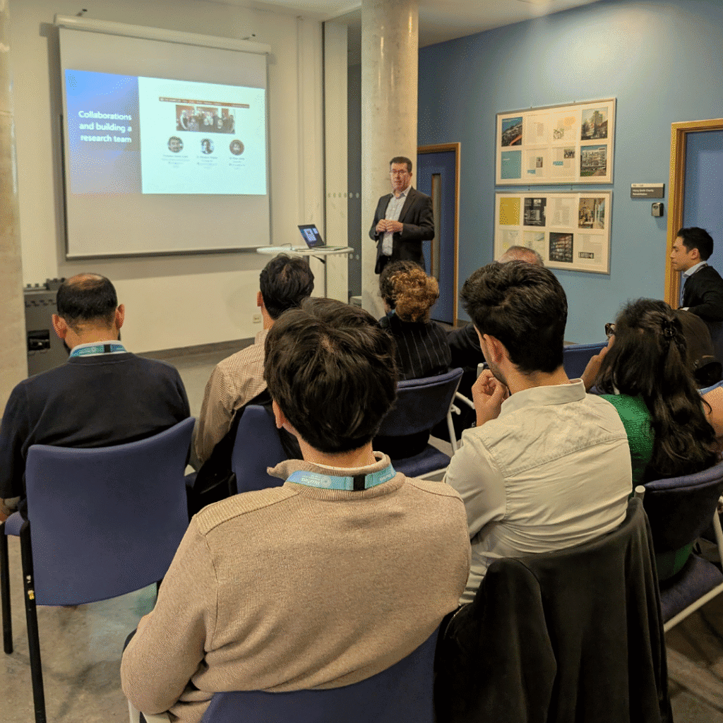 A speaker stands at the front of a small conference room giving a presentation . Several attendees are seated facing the screen, listening attentively. The room has blue walls, a door to the right, and posters displayed on the wall.