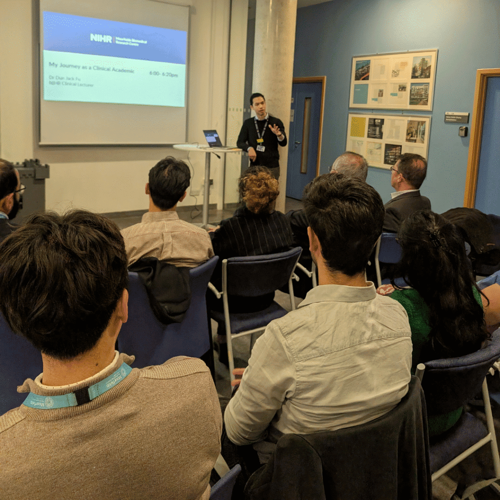 A speaker stands at the front of a small conference room giving a presentation . Several attendees are seated facing the screen, listening attentively. The room has blue walls, a door to the right, and posters displayed on the wall.