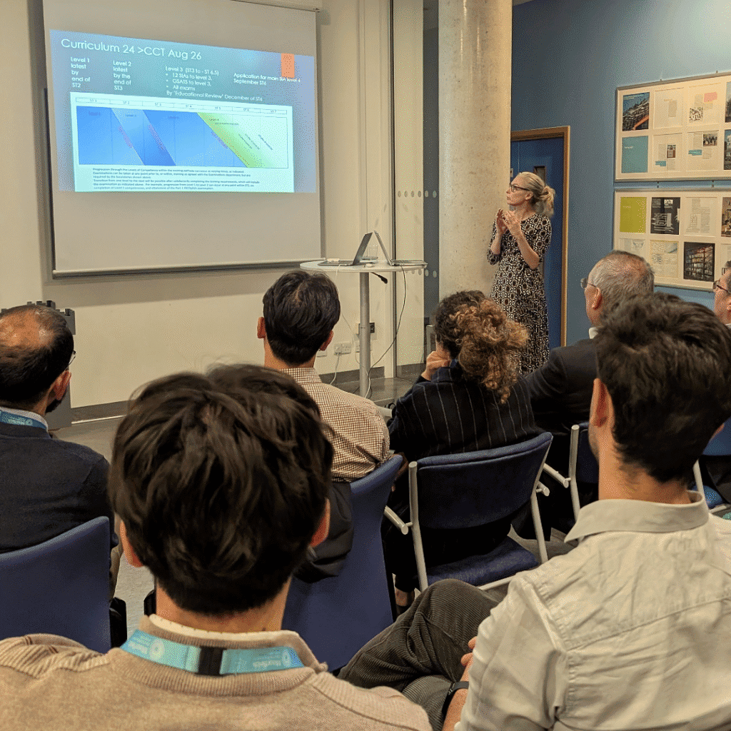 A speaker stands at the front of a small conference room giving a presentation . Several attendees are seated facing the screen, listening attentively. The room has blue walls, a door to the right, and posters displayed on the wall.
