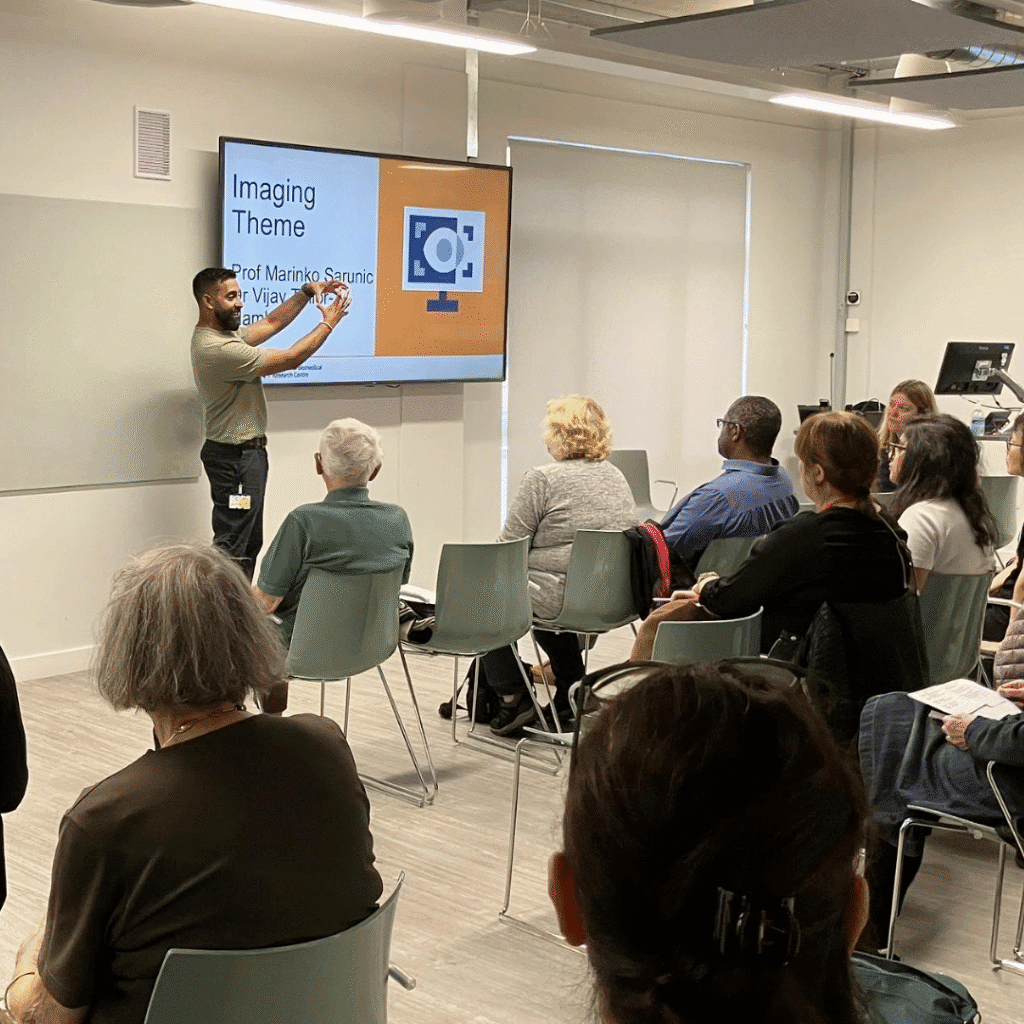 A speaker stands at the front of a room pointing to a large screen displaying a slide titled “Imaging Theme” with an orange and white graphic of a computer monitor and camera icon. Several attendees are seated in rows, attentively facing the presentation. The room has a bright, modern design with light-colored walls and flooring.