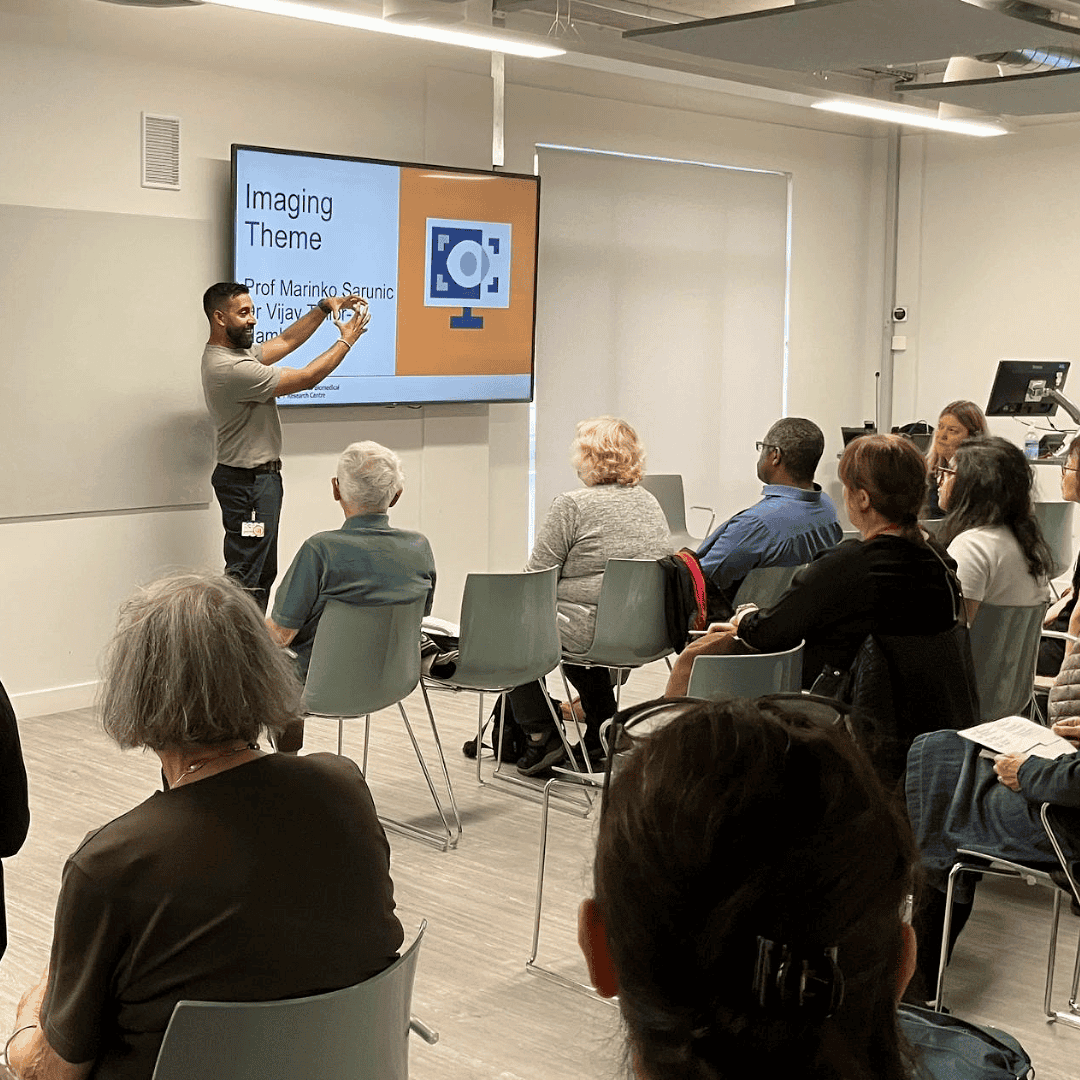 A speaker stands at the front of a room pointing to a large screen displaying a slide titled “Imaging Theme” with an orange and white graphic of a computer monitor and camera icon. Several attendees are seated in rows, attentively facing the presentation. The room has a bright, modern design with light-colored walls and flooring.