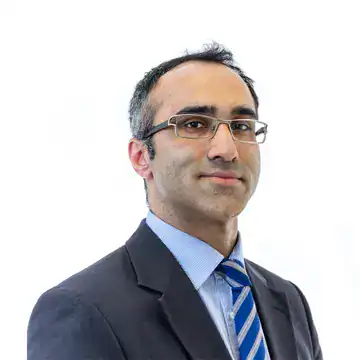 Professionally dressed man wearing a dark suit, light blue shirt, and blue striped tie, with rectangular eyeglasses and short dark hair graying at the temples, posed against a plain white background