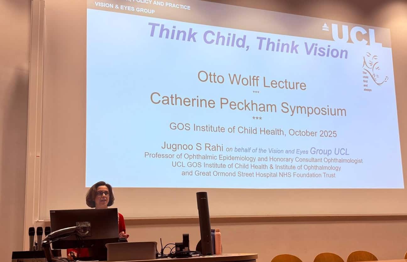 A woman stands at a podium in a lecture hall, delivering a presentation. She is speaking into a microphone with a laptop in front of her. Behind her, a large projection screen displays the title slide of her talk. The slide reads: “Think Child, Think Vision” and notes that the event is part of the Otto Wolff Lecture and Catherine Peckham Symposium, held at the GOS Institute of Child Health in October 2025. The speaker is Jugnoo S Rahi, representing the Vision and Eyes Group at UCL. Her titles include Professor of Ophthalmic Epidemiology and Honorary Consultant Ophthalmologist at UCL GOS Institute of Child Health & Institute of Ophthalmology and Great Ormond Street Hospital NHS Foundation Trust. The setting is formal and academic, with a focus on child health and vision research.