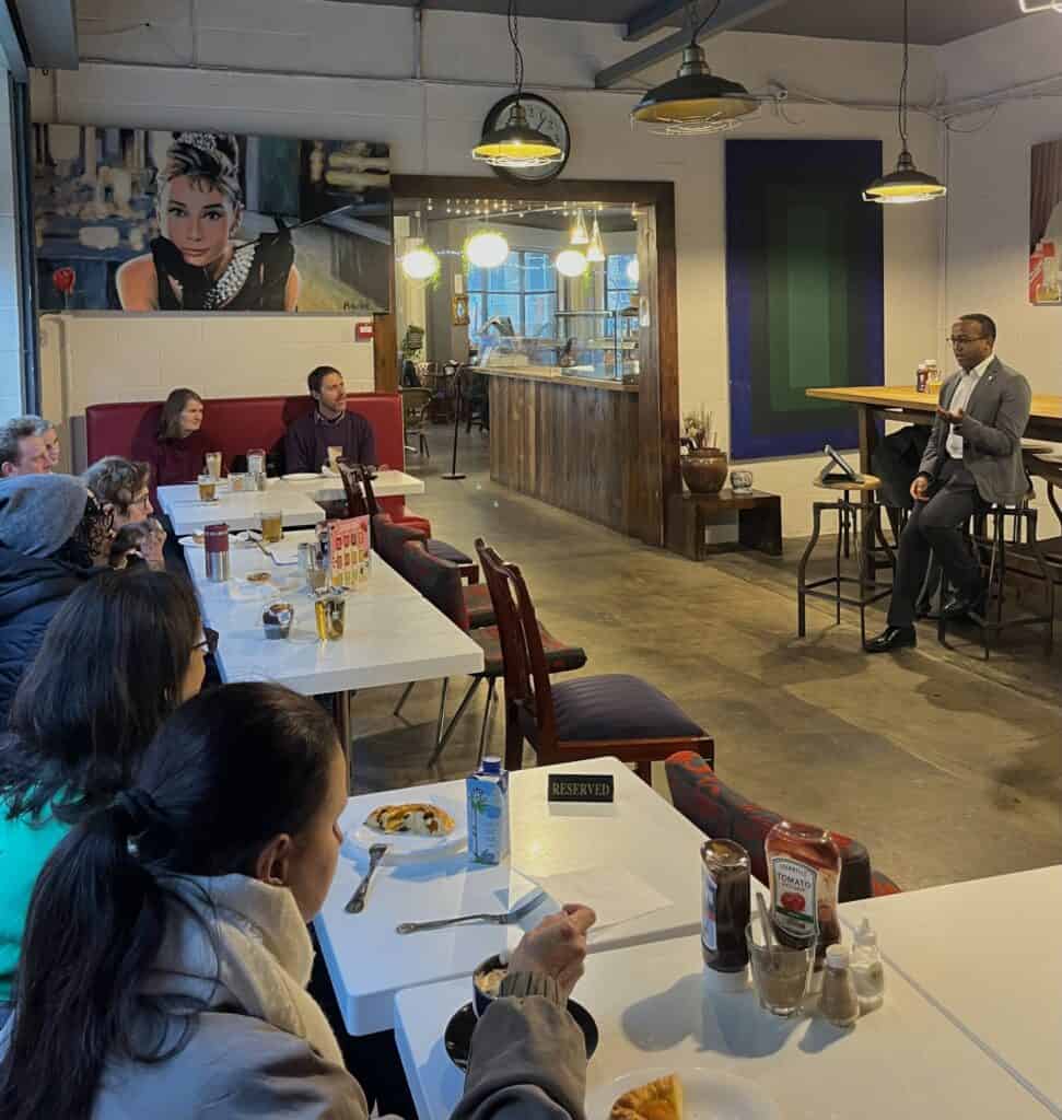 Dr Paul Nderitu is sat at the front of the coffee shop giving a talk to an audience of 8 attendees. Attendees are sat on booths facing Dr Nderitu and are engaging with the talk whilst enjoying coffee and pastries.