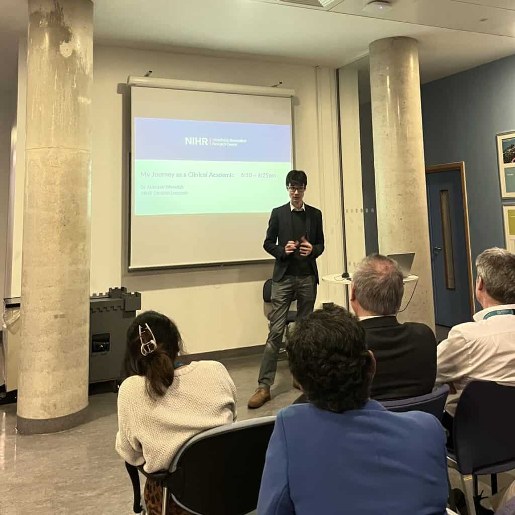 Dr Alasdair Warwick stood in front of an audience of clinical academics and trainees delivering a talk on his clinical academic journey. Behind him is a projector displaying the NIHR Moorfields Biomedical Research Centre logo.