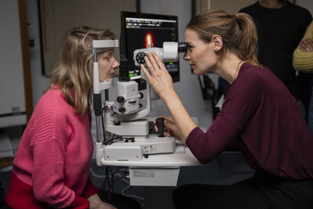 An ophthalmologist examines a patient’s eye using a specialist microscope in a clinical setting. There is diagnostic imaging displayed on a screen in the background.