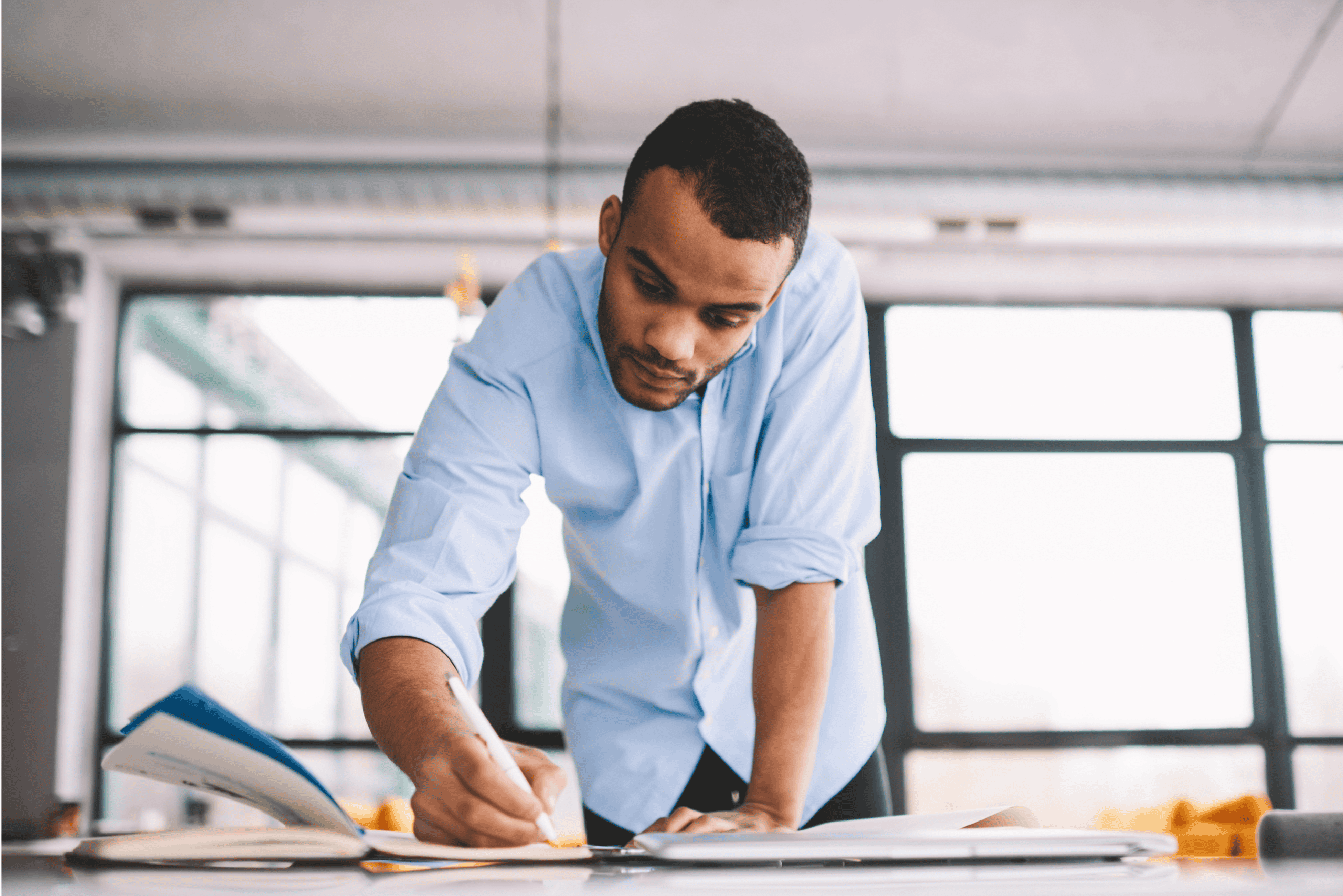 A stock image of a male person leans over a desk in a bright working space writing in a book.