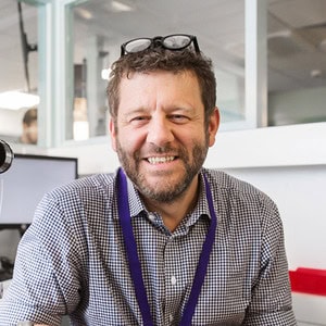 Image shows headshot of a white male with dark hair and greying beard, with glasses propped on his head and a broad smile