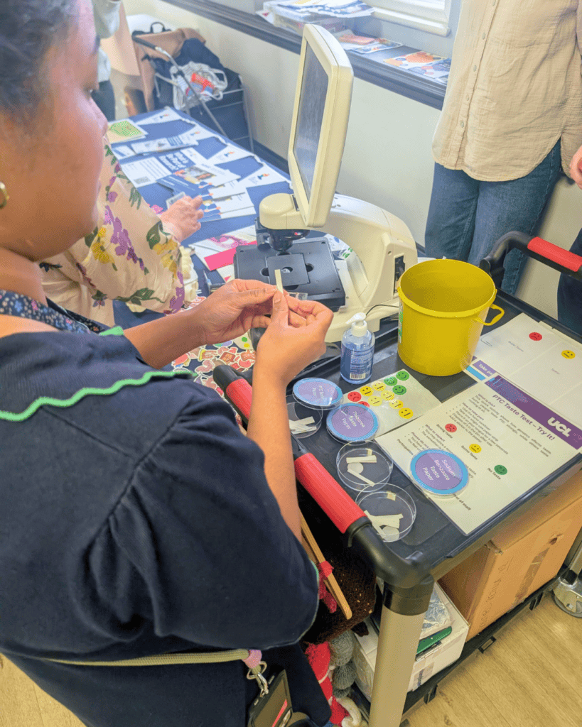 Close-up of a person using a microscope at an information stall, holding a prepared slide. The table displays petri dishes, small tools, stickers, and printed materials, alongside a yellow clinical waste container and hand sanitiser.