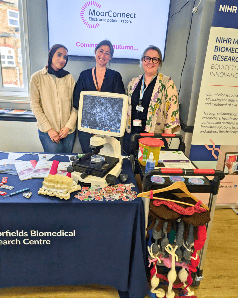 Three people standing behind a Moorfields Biomedical Research Centre display table with a microscope and educational materials. A screen behind them shows “MoorConnect electronic patient record – Coming autumn,” and a branded NIHR banner stands to the side.