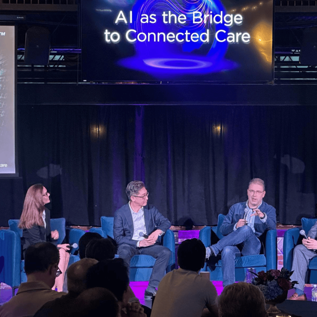 A panel discussion at a conference, with several speakers seated on a stage in front of a large audience. The speakers are sitting in blue chairs and holding microphones while engaging in discussion. A large screen above them displays the text “AI as the Bridge to Connected Care” with a stylised digital graphic. The stage is lit with purple lighting, and audience members are visible seated in the foreground.