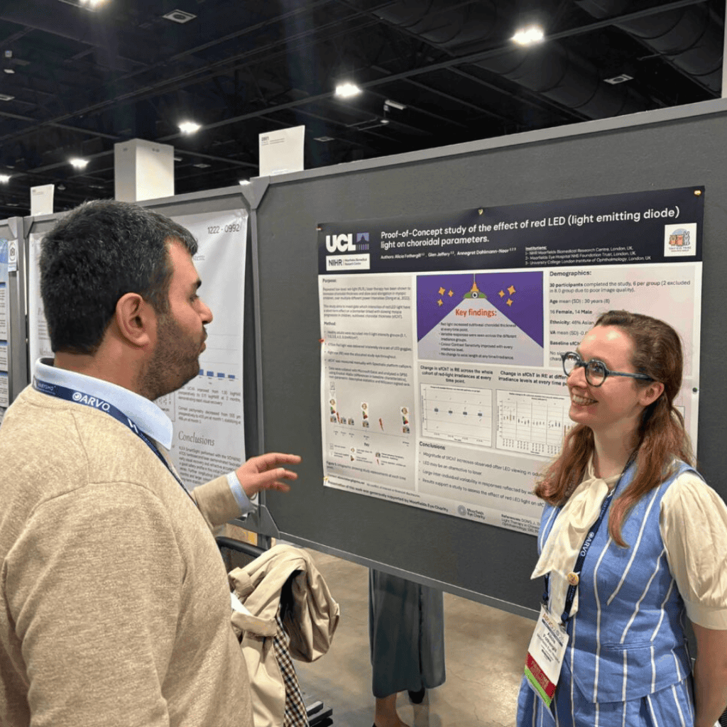 Two conference attendees stand in front of a research poster on a display board at an indoor exhibition hall. One person gestures toward the poster while speaking, and the other faces them, wearing glasses and a lanyard. The poster, branded with UCL and NIHR logos, is titled and includes sections such as key findings, demographics, charts, and diagrams. Attendees are visible in the background under bright overhead lighting.