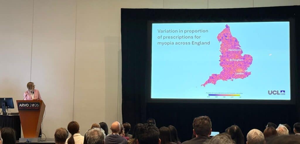 Image shows Annegret presenting at ARVO 20256. a woman is at a podia in a large room full of people and speaking about an image shows on a screen. The screen has a purple and pink coloured map of Englad and says Variation in proportion of prescriptions for myopia across England