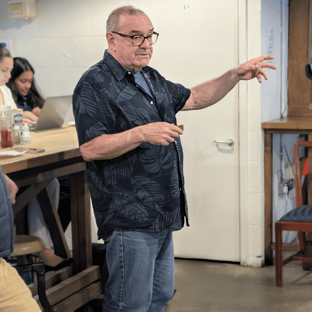 Prof Glen Jeffery is stood in a coffee shop gesturing with one hand as he gives a talk. He is wearing glasses, jeans and a blue shirt.