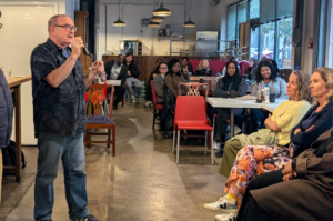Prof Glen Jeffery is standing and speaking to an audience in a coffee shop. The audience is seated audience arranged around tables. The room has large windows and overhead lights.