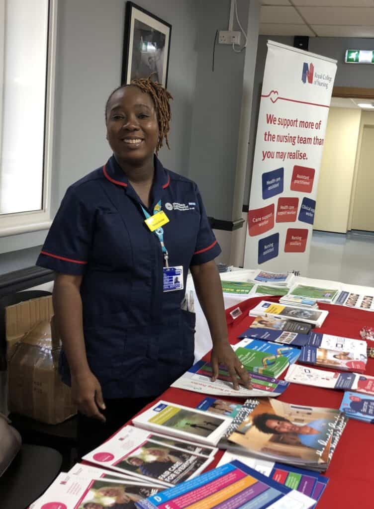 Nicola is in an NHS clinical uniform standing behind a table covered with leaflets at an information stand, with a Royal College of Nursing banner visible in the background.