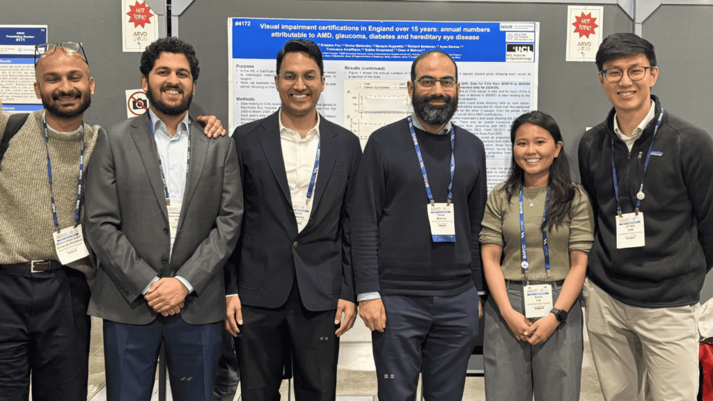 Six people stand side by side in front of a scientific poster presentation at a conference, each wearing name badges and conference lanyards. The poster behind them, titled about visual impairment certifications in England, includes charts and text, with “Hot Topic ARVO 2026” signs displayed on the wall nearby.