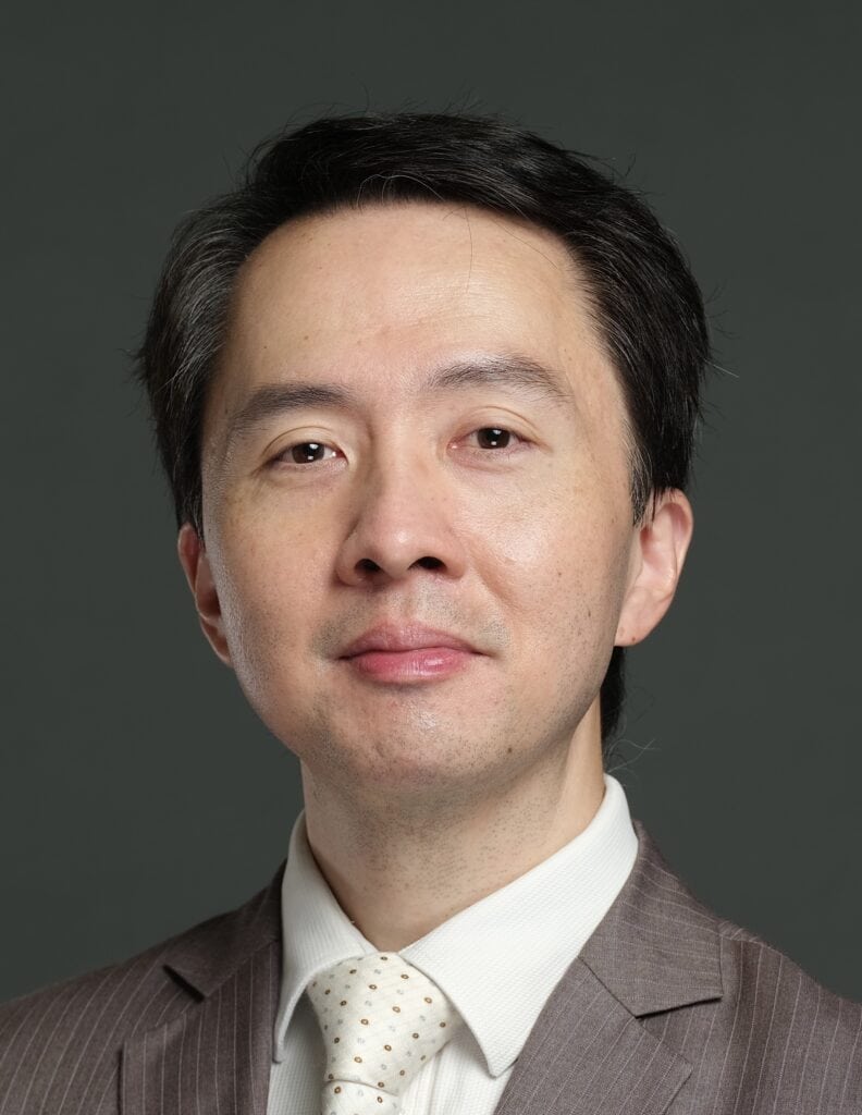 Head-and-shoulders portrait of Prof Patrick Yu Wai Man in a suit and tie, facing the camera against a plain, dark green background.