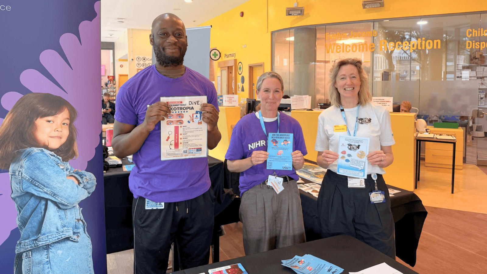 Three people standing behind an information table at a Moorfields Biomedical Research Centre event, each holding informational leaflets. A hospital reception area is visible in the background, with a sign reading “Welcome Reception,” and display materials arranged on the table.
