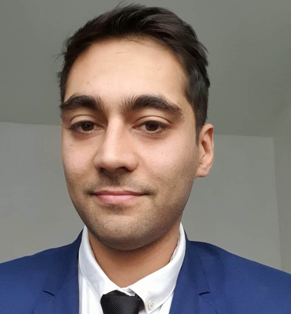 Headshot of Dr Shafi Balal wearing a navy suit jacket, white shirt, and black tie. He has short dark hair and is positioned against a plain, light-coloured indoor background.
