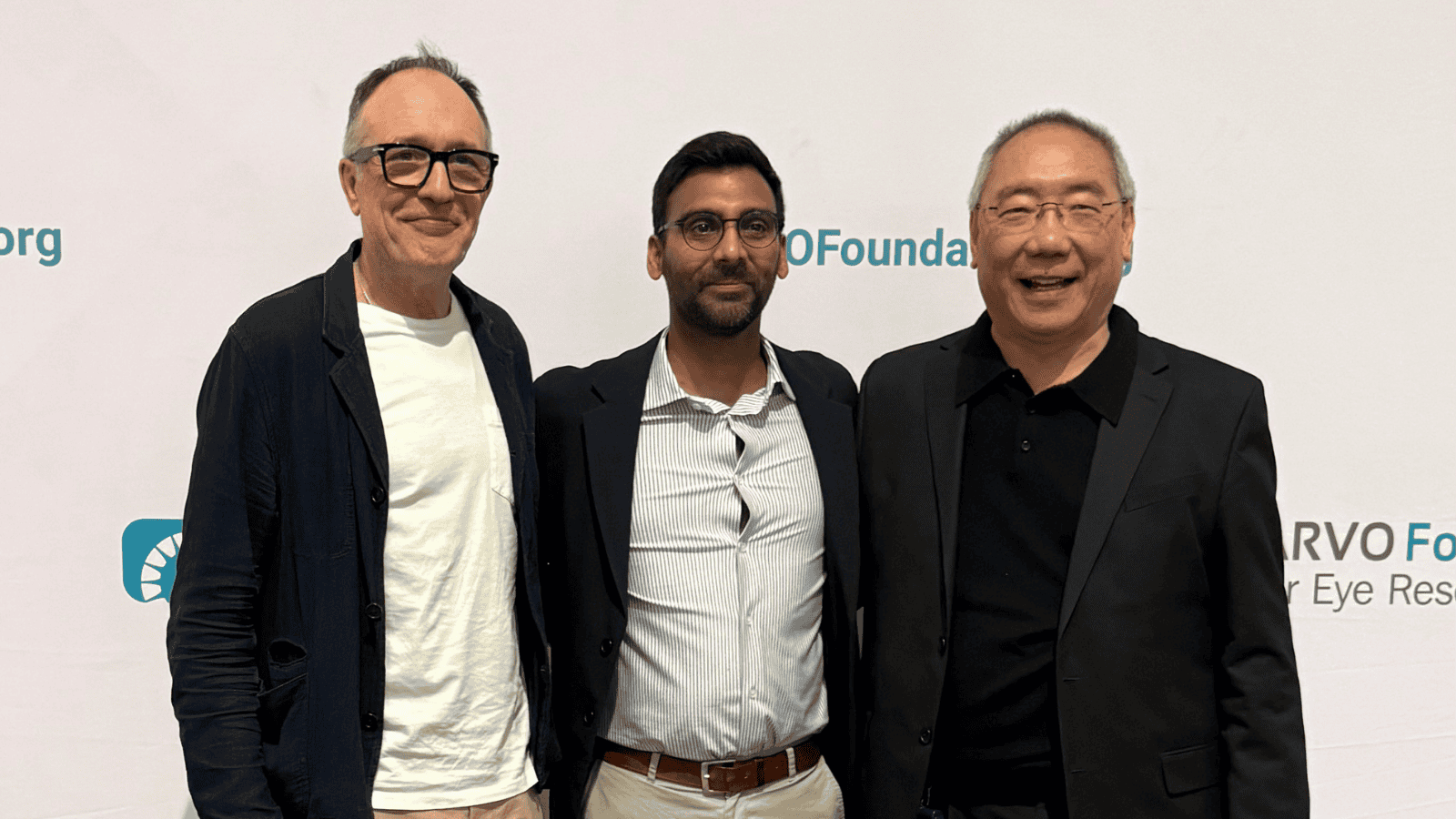 Three people stand side by side in front of a white conference backdrop with ARVO Foundation branding. They are dressed in smart-casual clothing, with jackets and shirts, and are posing for a photo. The background includes partial event logos and text, indicating a formal conference or awards setting.