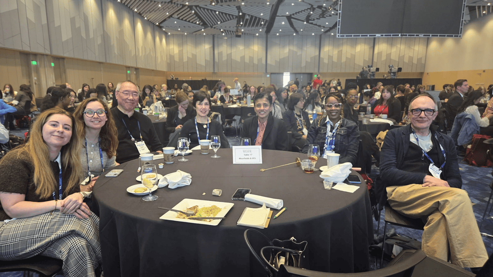A group of conference attendees seated around a round table at a large indoor event space. The table is set with drinks, plates, and notebooks, and a sign reading “WEAVER Table 17 Moorfields & UCL” is placed at the centre. The attendees face the camera, while many other tables and participants fill the background in a spacious, well-lit conference hall with a patterned ceiling and large screen displays.