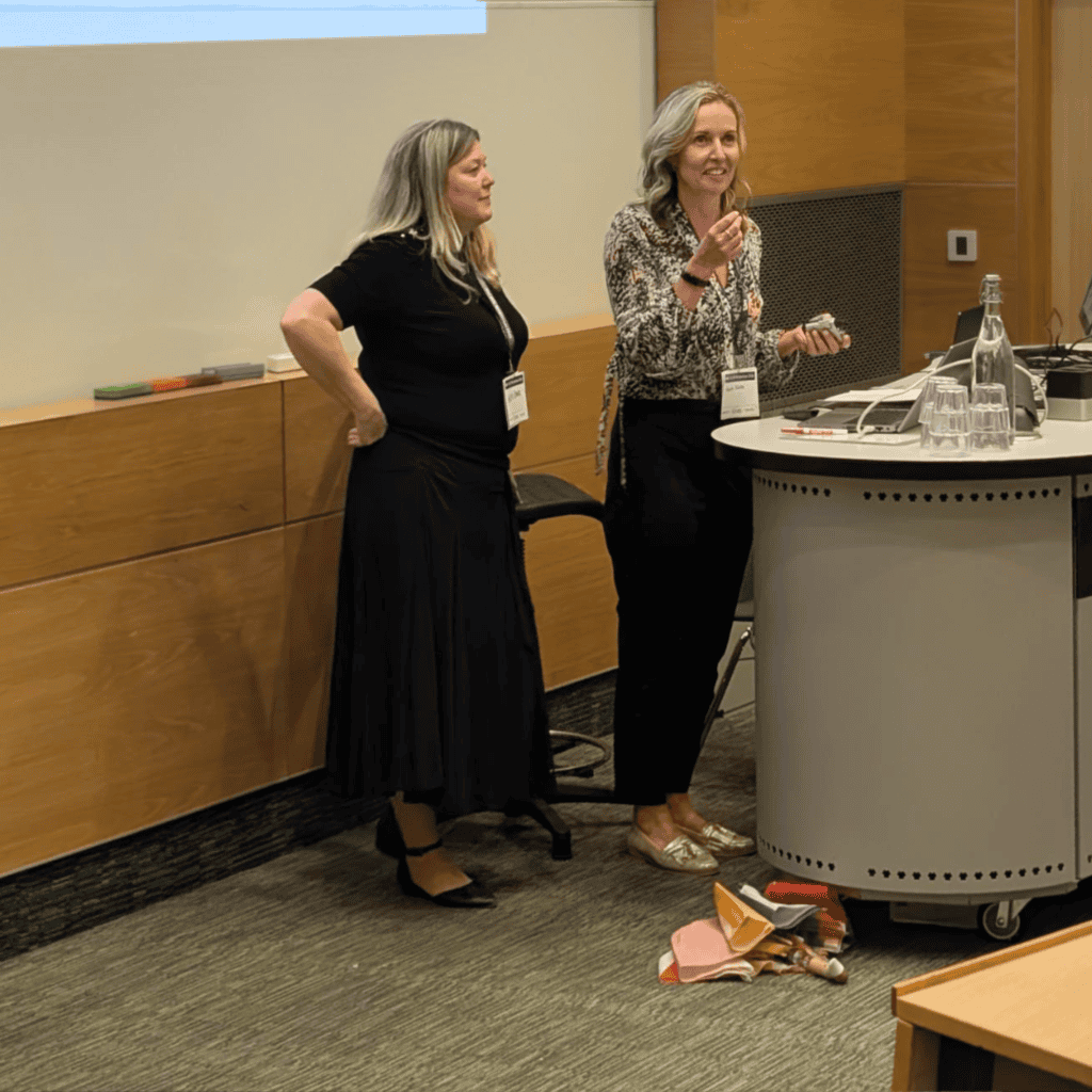 Two presenters stand at the front of a lecture theatre beside a podium with a computer and microphone, addressing an audience. A projected slide with bullet point text is displayed behind them on a large screen. One presenter is speaking while holding a small device, and the other stands nearby. On the desk are glasses, a water bottle, and presentation equipment, with wood-panelled walls and a door visible in the background.