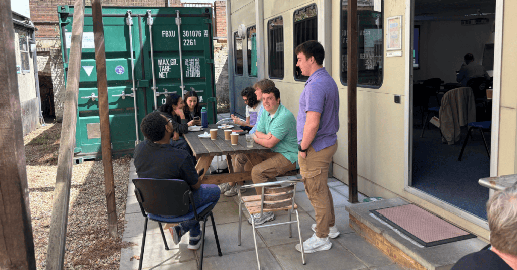 A small group gathered around a wooden picnic table in an outdoor courtyard, with drinks and plates on the table, a green shipping container in the background, and a building with windows and a door nearby.