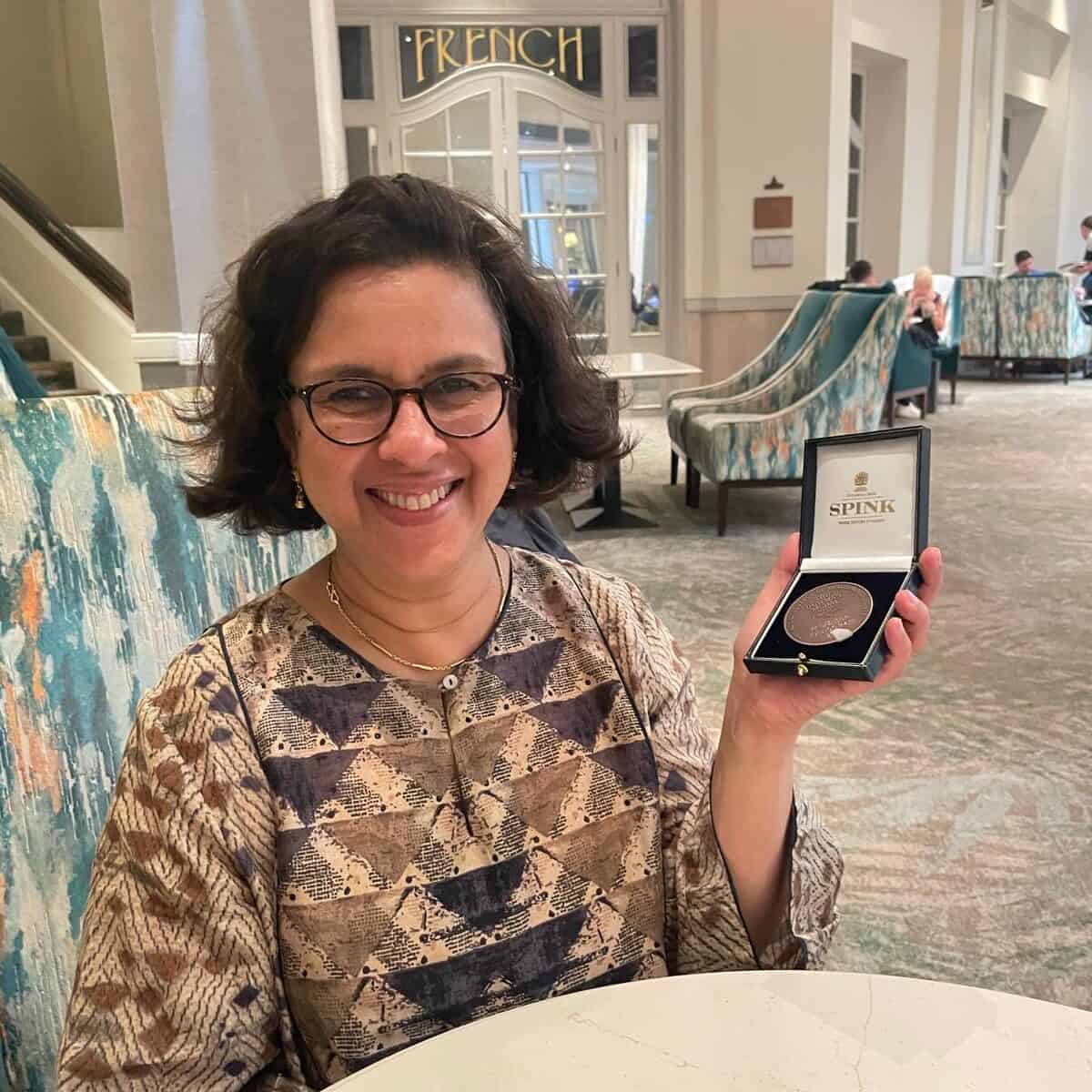 Prof. Jugnoo Rahi is seated at a table smiling at the camera and holding an open presentation case containing the Bowman medal.