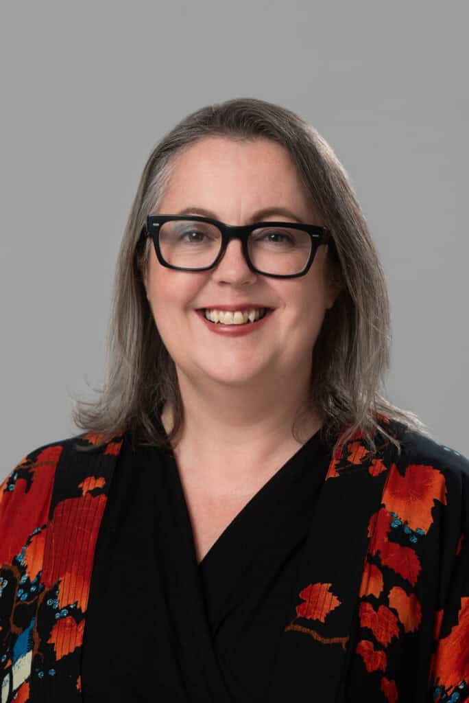 Image shows Amanda Carr a white woman with dark rimmed glasses and shoulder length hair wearing a red and black top