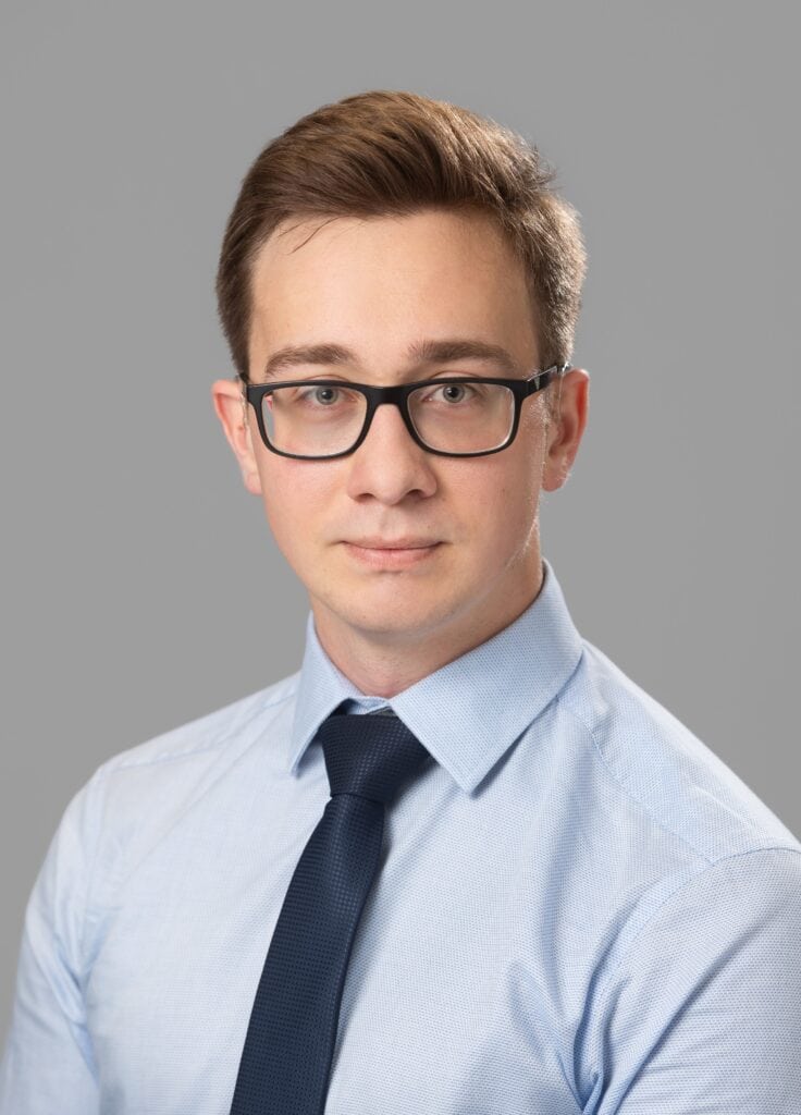 headshot of a person with black glasses, blue shirt and a black tie