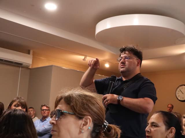 A person stands among a seated audience in an indoor meeting room, holding a microphone and gesturing with one hand as if speaking or asking a question. The person wears a dark short-sleeve shirt, glasses, and a wristwatch. Several other attendees sit nearby, some facing forward and others turned slightly toward the speaker. The room has warm lighting, beige walls, a visible air conditioning unit, a wall clock, and a large circular ceiling light fixture.