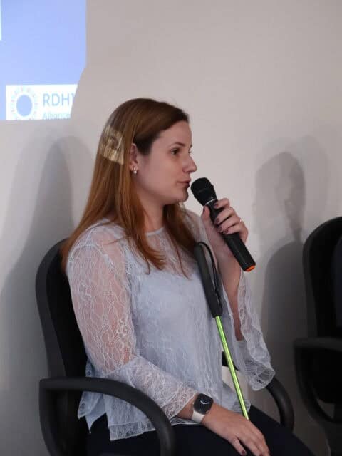 A person seated indoors holds a microphone close to their mouth while leaning slightly forward. The person wears a white lace long-sleeve top, dark trousers, and a smartwatch, and has long hair draped over one shoulder. The person is seated on a dark chair, and a green cane or stick rests diagonally across the lap. In the background, a plain wall displays a projected slide with partial visible text reading “RDH12,” indicating a presentation setting.