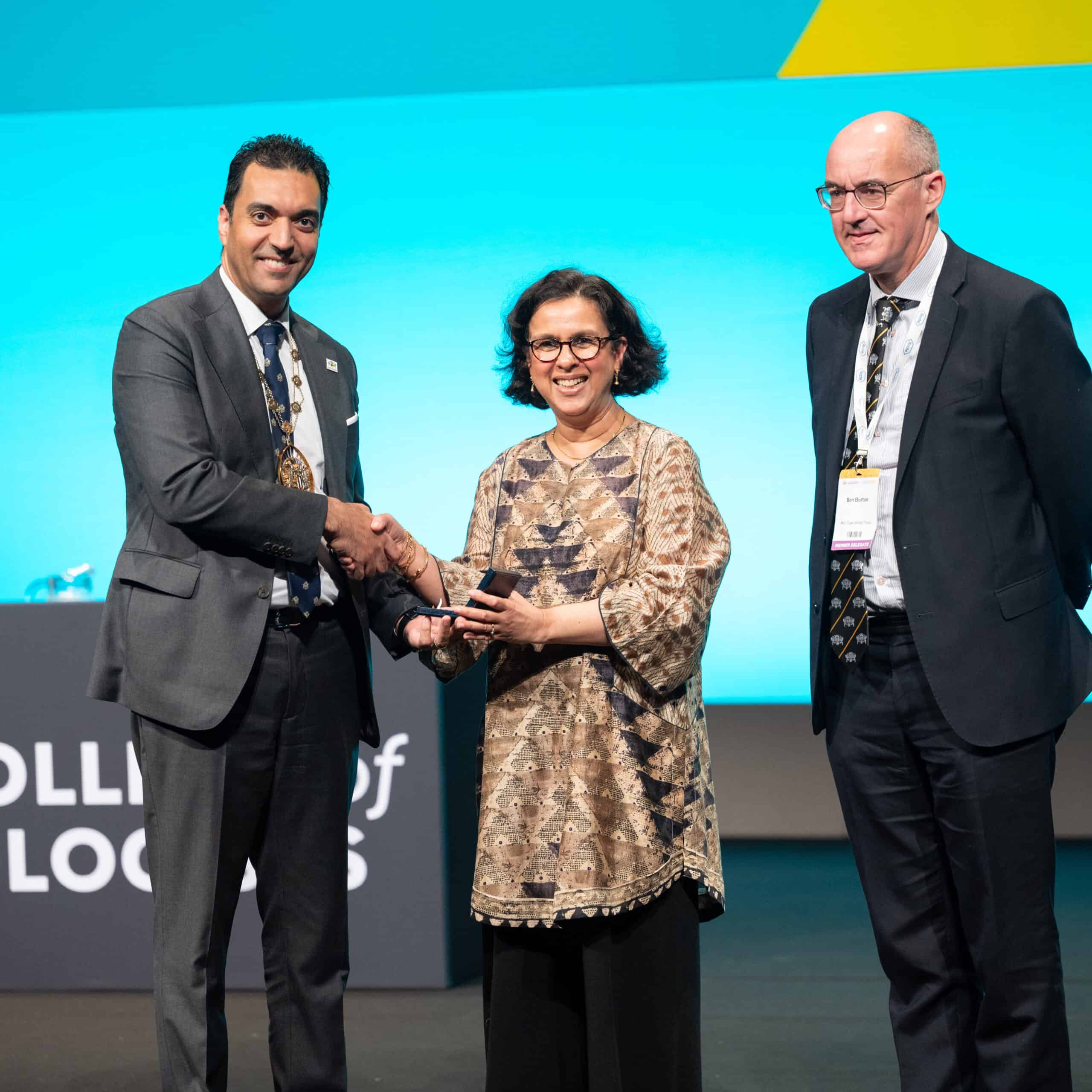 Jugnoo is on a stage being presented with the Bowman medal by two men. One is presenting her with the medal while shaking hands. The backdrop is brightly lit and includes a podium at the front.
