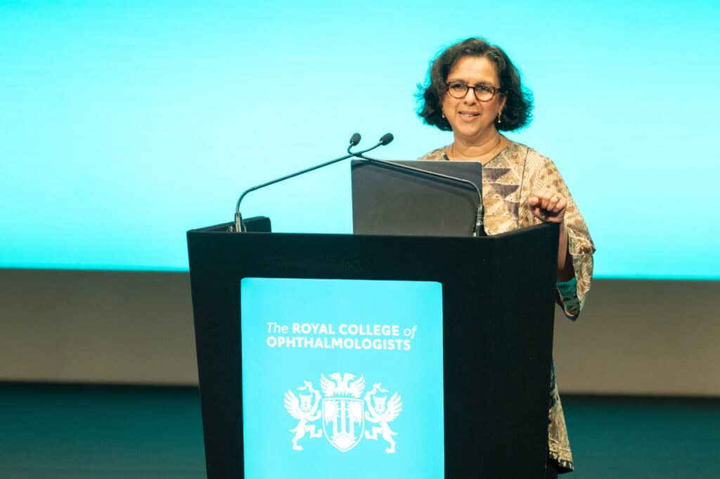 Prof. Jugnoo Rahi stands at a lectern delivering a speech, with microphones and a laptop in front. The lectern displays “The Royal College of Ophthalmologists,” and the background is a lit conference stage.