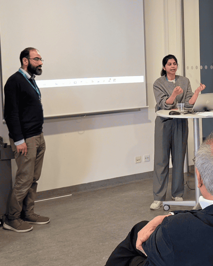 Two presenters stand at the front of a meeting room beside a projection screen and a small table with a laptop.
Audience members are seated in the foreground, indicating a live presentation setting.