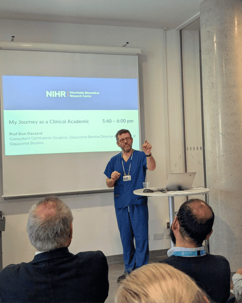 A speaker presents in front of a slide reading “NIHR | Moorfields Biomedical Research Centre” and “My Journey as a Clinical Academic.”
Audience members face forward while the speaker gestures beside a laptop and lectern table.