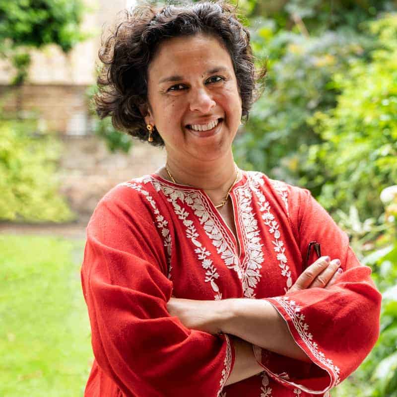 Prof. Jugnoo Rahi standing outdoors with her arms folded, wearing an orange top with embroidered detailing and smiling at the camera.