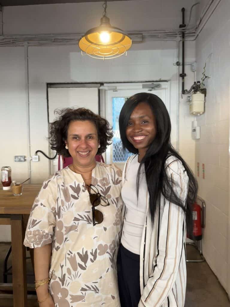 Prof Jugnoo Rahi and Ruke Ejegreh standing side by side, both smiling at the camera. The background shows a café with wooden tables, stools and overhead lighting.