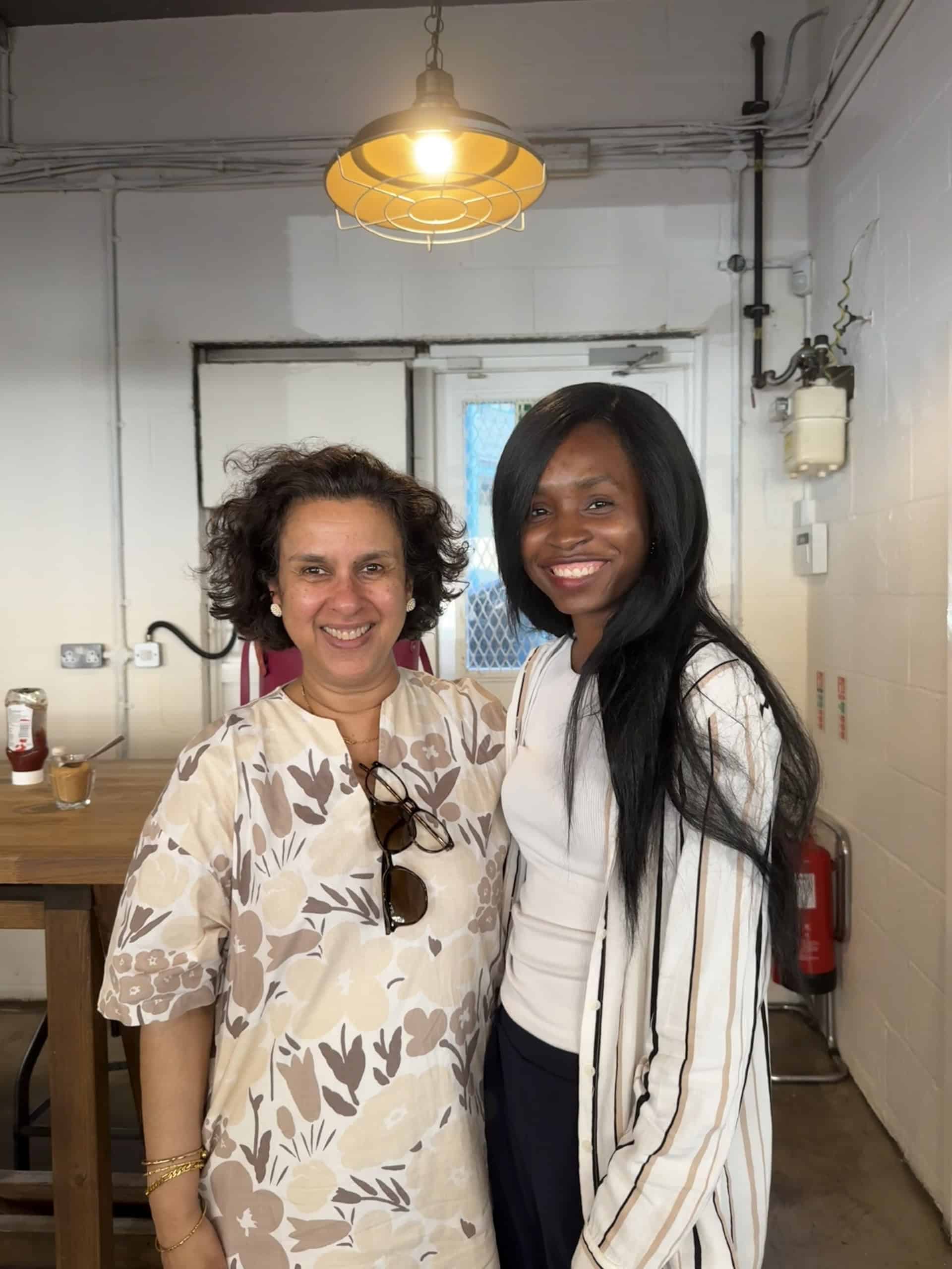 Prof Jugnoo Rahi and Ruke Ejegreh standing side by side, both smiling at the camera. The background shows a café with wooden tables, stools and overhead lighting.