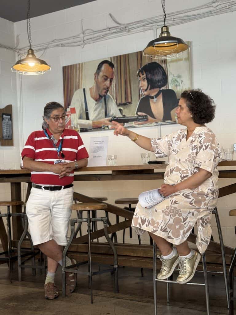 Prof Jugnoo Rahi is seated on a high stool holding notes and gesturing while speaking, with Dr Subhojit Chakraborty stood next to her listening. They are in a café with wooden tables, stools, hanging lights, with a large painting on the wall behind them.